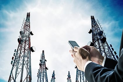 Torres de telecomunicação vistas de baixo contra o céu, com uma mão segurando um celular em primeiro plano